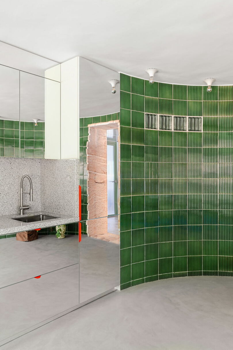 A modern kitchen with green vertical tile walls, a mirrored cabinet, a stainless steel sink, and a speckled countertop.