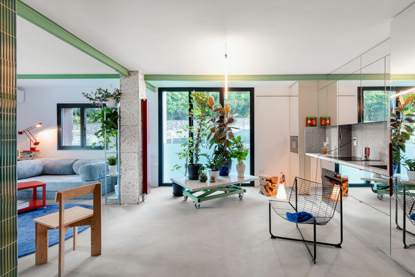 Modern living room with a mix of furniture including a blue sofa, wooden chair, and wire chair. Indoor plants decorate the space, and large windows provide natural light.