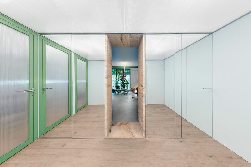 A modern hallway with green-framed glass doors on the sides, light wood flooring, and a central opening leading to a room with plants.