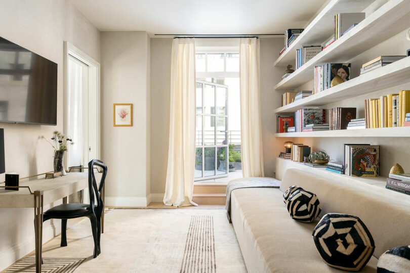 A modern living room featuring an Elizabeth Graziolo-inspired beige sofa, open shelves adorned with books, a small desk with a chair, and a wall-mounted TV. Sunlight gracefully streams through a window draped in light curtains.