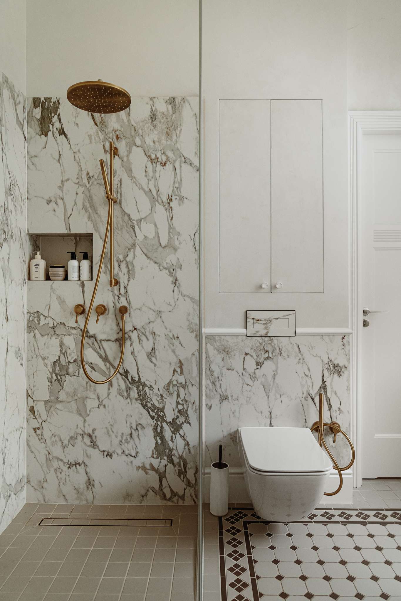 In the shower, bold marble continues alongside minimalist brass fittings and a built-in niche. Every surface, from the patterned floor tiles to the custom lighting, feels carefully considered, creating a calm and elegant retreat.