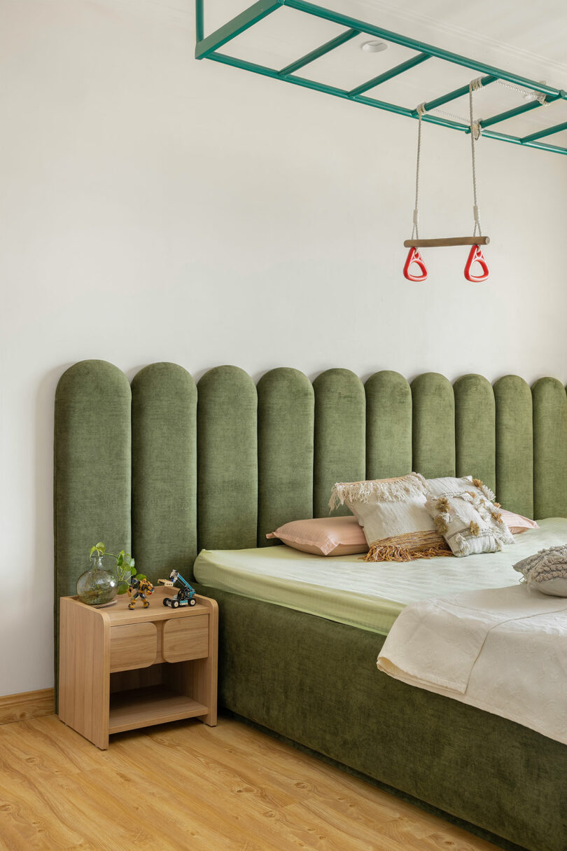 A bedroom with a green upholstered bed, light bedding, a wooden nightstand with toys and plants, and gym rings hanging from a green ceiling structure.