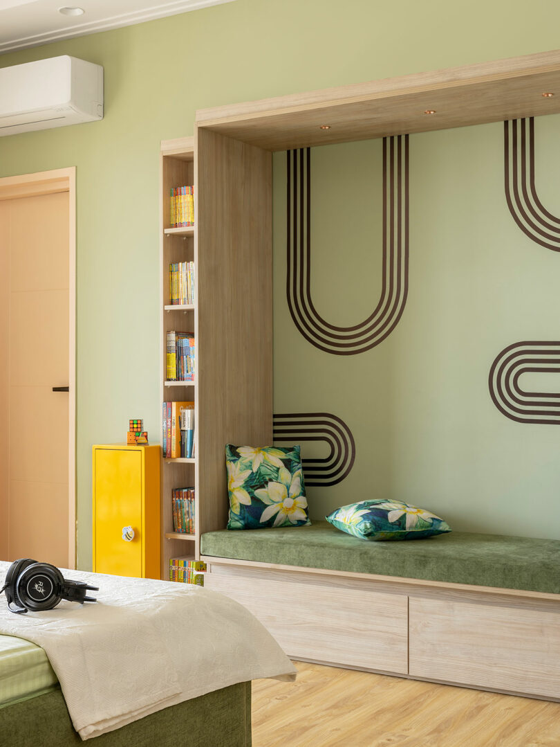 A cozy reading nook with floral cushions, built-in shelves filled with books, a yellow cabinet, and geometric wall patterns in a light green bedroom.