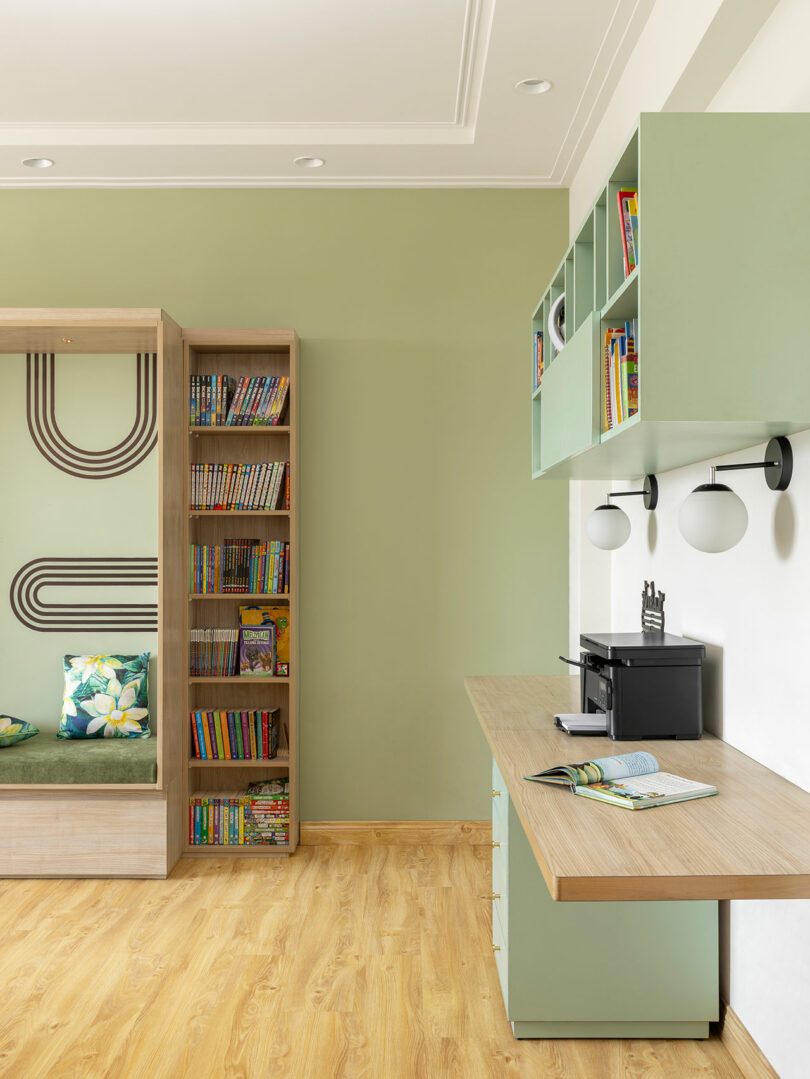 Modern study room with green walls, a wooden desk, open shelves with books, a bench with cushions, and a light wood floor.