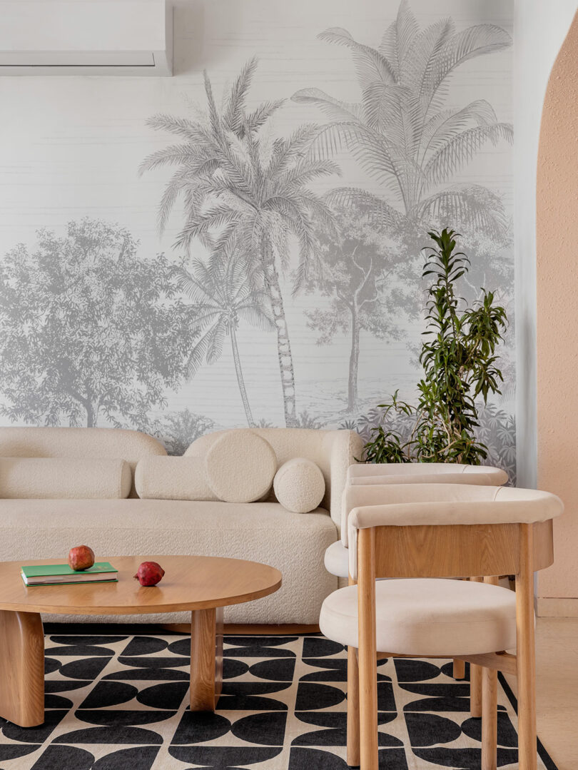 A modern living room with a cream sofa, a wooden coffee table with books and apples, a patterned rug, and a plant, set against a wall with a grayscale palm tree mural.