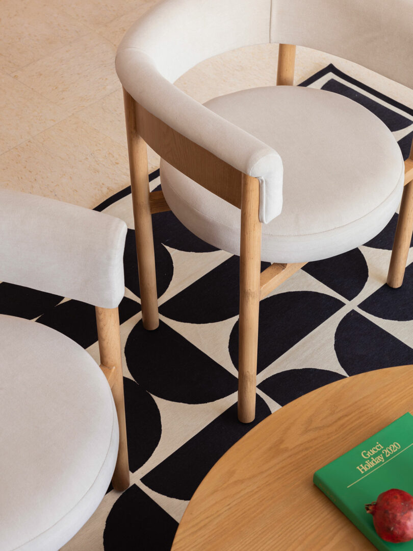 Two modern chairs with light upholstery and wooden legs are placed on a black and white geometric rug next to a wooden table with a green book and a red fruit.